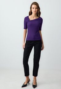 Top aderente viola a coste con maniche corte e un collo asimmetrico, caratterizzato da un bottone decorativo. Abbinato a pantaloni neri slim e tacchi.