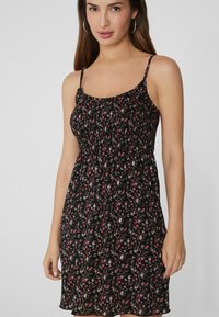 Schwarzes Blumenkleid mit dünnen Trägern, das einen gerafften Oberteil und ein fließendes Design aufweist. Kleine pinke und weiße Blumen im gesamten Stoff.
