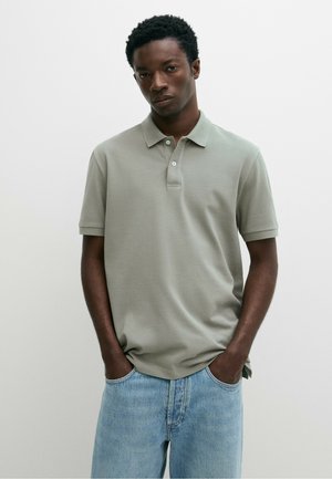Junger Mann mit kurzen lockigen Haaren, trägt ein hellgraues Poloshirt und blaue Jeans, steht mit den Händen in den Taschen vor einem schlichten Hintergrund.
