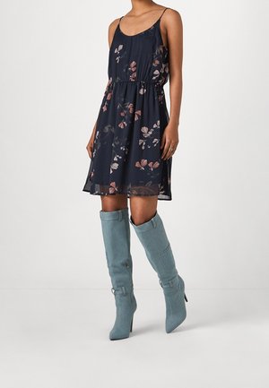 Frau trägt ein ärmelloses marineblaues Kleid mit pinken Blumenmustern und hellblauen kniehohen Wildlederstiefeln mit hohen Absätzen, steht vor einem einfarbigen Hintergrund.