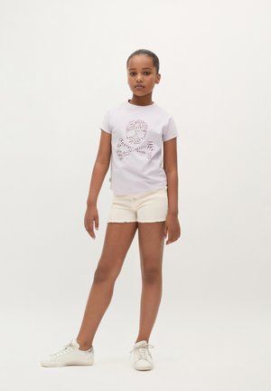 Meisje die staat en een lichtpaarse t-shirt met doodshoofdprint draagt, crèmekleurige shorts en witte sneakers, tegen een effen witte achtergrond.