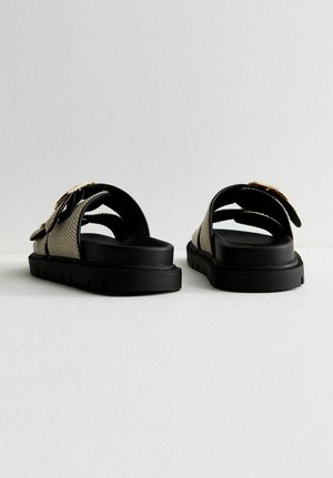 Sandalias slip-on en negro y beige con una correa texturizada y suela de goma gruesa, con un acento dorado en los detalles de la hebilla.