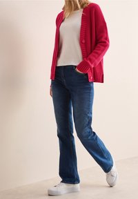 Punainen ribbetty neulejakku napilla, yhdistetty valkoiseen t-paitaan ja tummansinisiin farkkuihin. Valkoiset lenkkarit täydentävät asua.