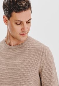 Beżowy sweter o teksturze, z okrągłym dekoltem, z wzorem w plaster miodu. Materiał wydaje się miękki i przewiewny.