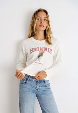Jonge vrouw met blond haar draagt een witte trui met tekst "BROWNIE" en een afbeelding van een teddybeer, gecombineerd met lichtblauwe spijkerbroek, staand tegen een witte muur.
