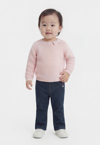 Kleinkind steht, trägt einen hellrosa gestrickten Pullover, dunkelblaue Hose und weiße Schuhe vor einem schlichten weißen Hintergrund.