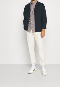 Giacca blu marina sopra una camicia a quadri rossi e bianchi, abbinata a pantaloni color crema e sneakers bianche con dettagli rossi.