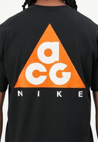 Schwarzes Kurzarm-T-Shirt mit einem großen orangefarbenen Dreieck, auf dem "acg" in Weiß und "NIKE" in Weiß darunter steht. Aus Baumwollmaterial.