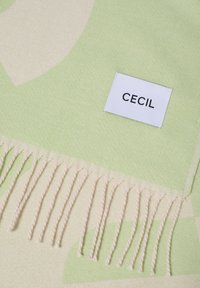 Grøn og cremefarget vævet tæppe med frynsekant; har et geometrisk mønster. En mærkat med "CECI" er syet fast på stoffet.