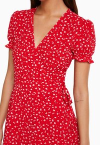 Vestido floral rojo con escote en V, mangas cortas abullonadas y un lazo en la cintura. La tela tiene un aspecto texturizado con pequeñas flores blancas.