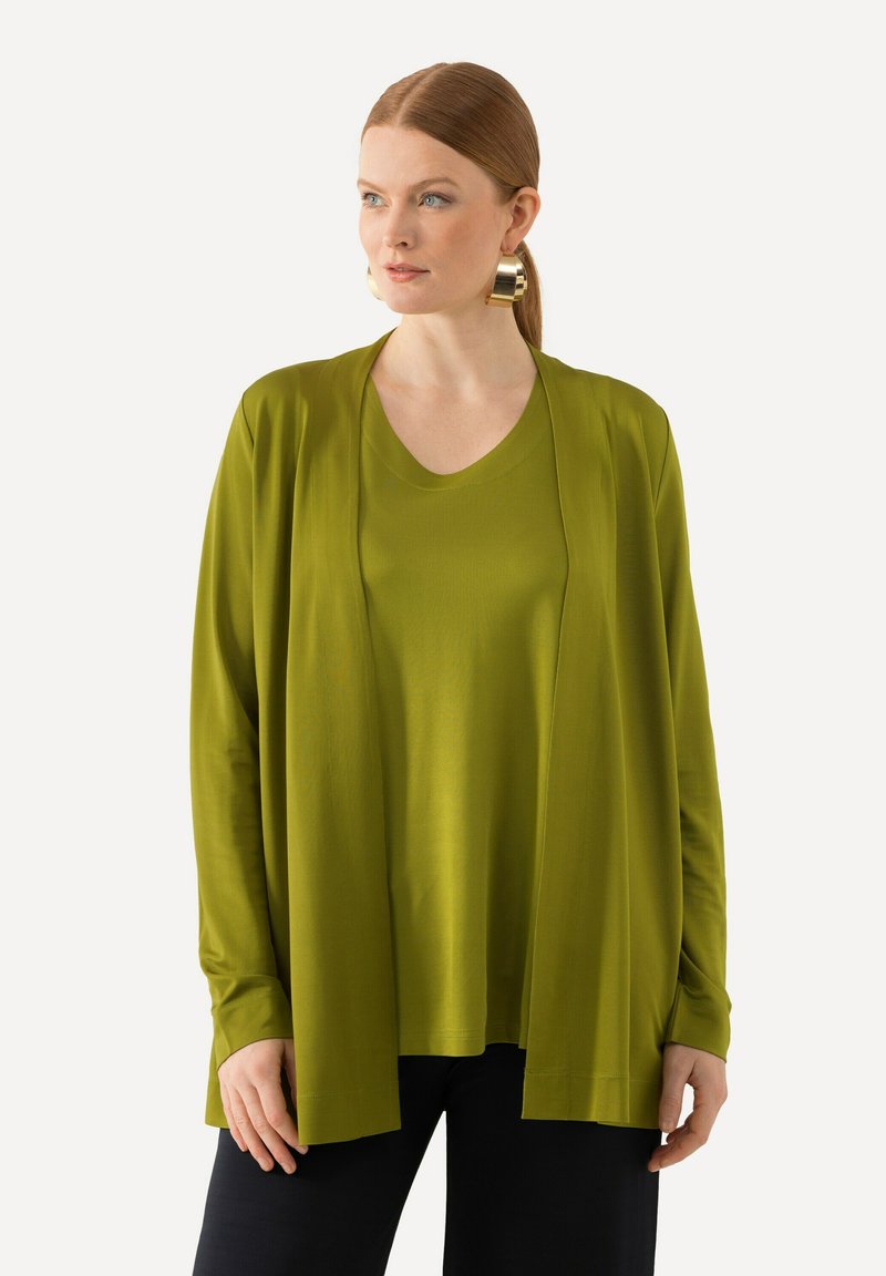 Olivgrüner, leichter Cardigan, über einem passenden T-Shirt getragen, kombiniert mit dunklen Hosen. Glatte Textur, lässiges Design, offenes Vorderteil.