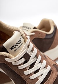 Brown suede sneakers with cream laces, featuring a textured surface and a prominent logo on the tongue. Accents include a beige midsole.