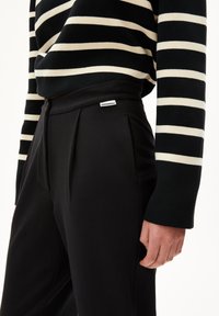 Personne portant un pantalon taille haute noir avec une petite étiquette "ARMEDANGELS" et un haut à manches longues noir et blanc à rayures.