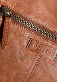 Barna bőrből készült kiegészítő, texturált felülettel, sárgaréz cipzárral és varrott címkével, amelyen a "DEPECHE" felirat található.