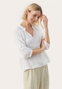 Balta lino bluzelė su laisvu kirpimu, V formos iškirpte ir raukšlėtu apykakliu. Yra trijų ketvirčių pūstučių rankovės ir minkšta, tekstūruota medžiaga.