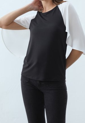 Vrouw draagt een zwarte top met witte, wapperende mouwen en een zwarte spijkerbroek, poserend met één hand achter haar hoofd en de andere op haar heup.