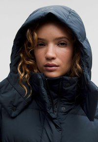 Veste matelassée navy avec un col haut et une capuche réglable. Présente des détails couturés et une fermeture éclair. Texture lisse et matelassée.