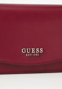Portafoglio in pelle rossa con un design elegante, con bordi arrotondati e il distintivo dorato "GUESS EST 1981". Texture liscia con cuciture visibili.