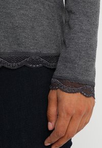 Närbild av en hand bredvid en grå ribbad tröja med lång ärm och svart spetskant vid ärmslut och nederkant, bärs över mörka byxor.