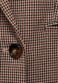 Gros plan sur un tissu à motif pied-de-poule bordeaux, noir et beige, avec un bouton brun foncé et une petite poche passepoilée sur une veste.