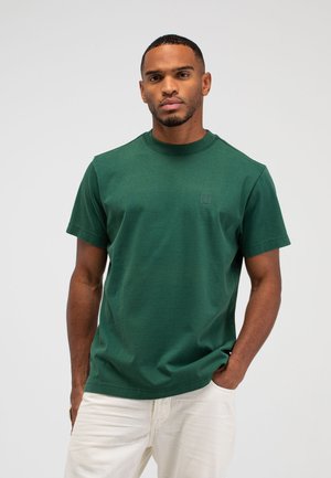 Man die een bosgroen T-shirt met korte mouwen en crèmekleurige broek draagt, staat met één hand in de zak tegen een effen lichte achtergrond.