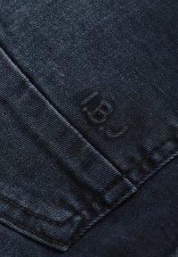 Mörkt blå denimtyg med en strukturerad yta och en subtil präglad logotyp. Sömmen är sydd med kontrasterande tråd.