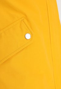 Yellow fabric close-up featuring a patch pocket with silver hardware and visible stitching details. Smooth texture with matte finish.