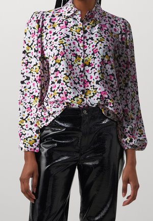Langærmet skjorte med knapper og blomsterprint i pink, hvidt og gult, stoppet ned i sorte, blanke bukser med tekstur.