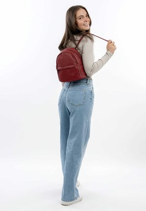 Roter Leder-Rucksack mit runder Form, ausgestattet mit einer Vorderseite-Tasche mit Reißverschluss und verstellbaren Trägern. Lässig zu hellblauen Jeans getragen.