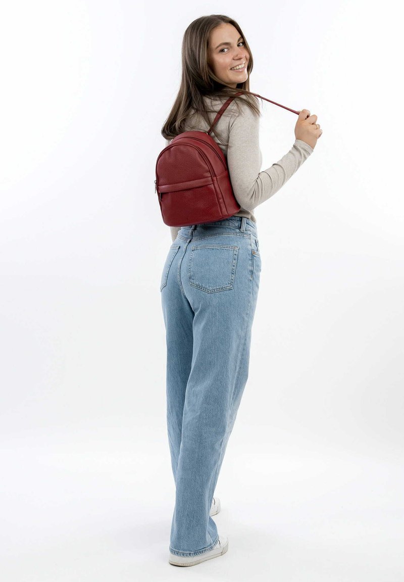 Zaino in cuoio rosso con forma arrotondata, dotato di una tasca frontale con cerniera e spallacci regolabili. Indossato in modo casual con jeans azzurri chiari.