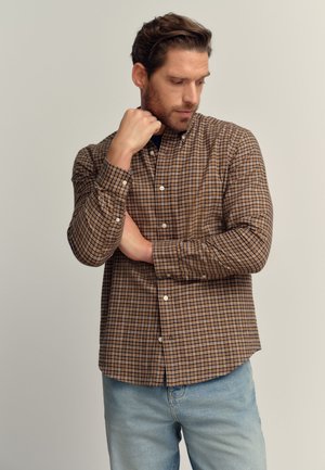Mann trägt braun kariertes Hemd zum Knöpfen und hellblaue Jeans, schaut nach unten, eine Hand berührt seinen Nacken, die andere ist verschränkt.