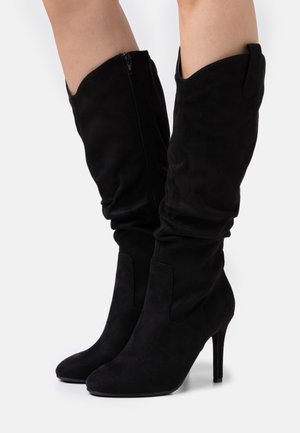 Botas de ante negras hasta la rodilla con punta afilada y tacón alto. Cuentan con un cierre de cremallera en el costado y un diseño holgado en la parte superior.