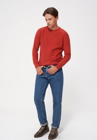 Czerwony sweter o teksturze, niebieskie dżinsy oraz brązowe buty na sznurówki. Ubranie charakteryzuje się luźnym krojem i podwiniętymi mankietami spodni.