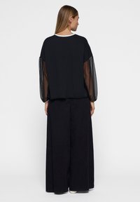 Zwarte top met doorzichtige lange mouwen, ronde halslijn en een losse pasvorm, gecombineerd met zwarte wijde broek. Minimalistisch ontwerp, geen zichtbare patronen.