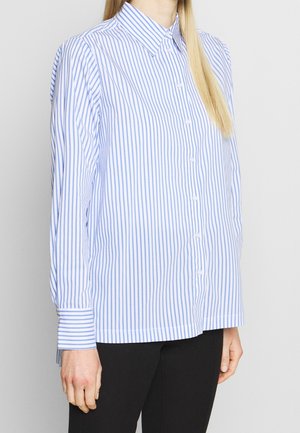 Vrouw draagt een blauwe en witte overhemd met verticale strepen, lange mouwen en een kraag, gecombineerd met een zwarte broek.