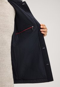 Manteau marine en tissu texturé, doté d'un intérieur matelassé et d'une poche zippée rouge. L'intérieur présente une finition lisse avec des détails de couture subtils.