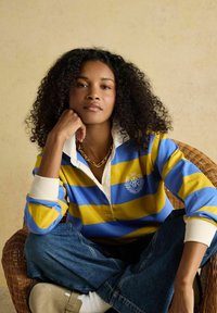 Femeie cu păr creț, purtând un tricou de rugby cu dungi albastre și galbene, blugi albaștri și un colier cu zale aurii, așezată pe un scaun de nuiele.