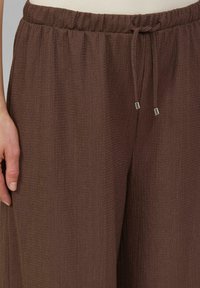 Hnědé široké kalhoty s texturou vyrobené z lehké látky, s elastickým páskem a stahovací šňůrkou s kovovými konci.