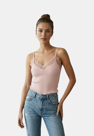 Donna con i capelli raccolti in uno chignon, che indossa una canottiera rosa bordata di pizzo e jeans blu, in piedi con una mano in tasca davanti a uno sfondo semplice.