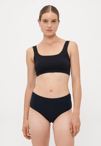 Woman wearing black high-waist briefs and a matching black crop top with lace trim on shoulder straps, standing against a white background.