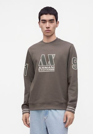 Młody mężczyzna ubrany w brązową bluzę Armani Exchange z logo i cyframi na rękawach, w połączeniu z jasnoniebieskimi dżinsami, stojący na białym tle.