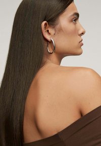 Vista lateral de una persona con cabello largo y lacio de color castaño, vistiendo una blusa marrón sin hombros y grandes aretes de plata en forma de aro. Se aprecia una textura de piel lisa.