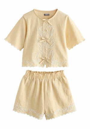 Jasnobeżowy komplet składający się z krótkiego topu i szortów z falowanymi brzegami i białym kwiatowym haftem, z przednimi kokardkami na topie.