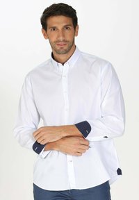 Chemise blanche à manches longues avec boutons, présentant des poignets à accents bleus, un col classique et une texture lisse, portée avec les bras croisés.