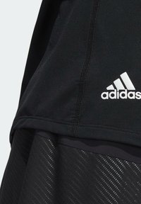 Gros plan sur un vêtement de sport noir Adidas montrant un ourlet cousu, un tissu texturé et le logo blanc Adidas sur le côté inférieur du vêtement.