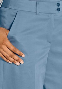 De hand van een persoon rust op de zak van lichtblauwe op maat gemaakte broek met dubbele knoopsluiting en riemlussen.