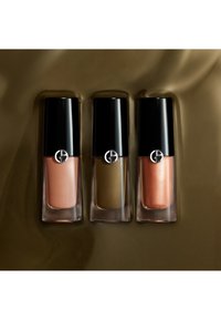 Tre flaconi di smalto Armani in tonalità nude, oliva e rame metallizzato con tappi neri su uno sfondo di raso marrone.