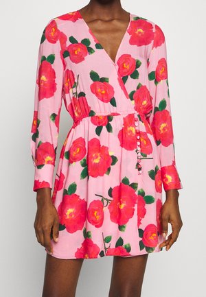 Femme portant une robe portefeuille rose avec des manches longues et de grandes imprimés floraux rouges et jaunes ainsi que des boutons sur le côté.