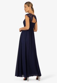 Abito lungo blu navy con maniche a cappuccio in pizzo e corpetto aderente, che si allarga dalla vita. Dettaglio in pizzo strutturato sulla schiena.