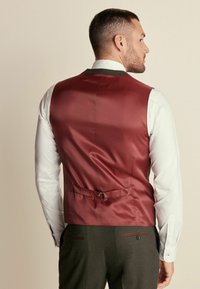 Bordo satenski prsluk s glatkom teksturom, s dizajnom koji se stegne straga i s detaljem srebrne kopče, nošen preko bijele košulje.
